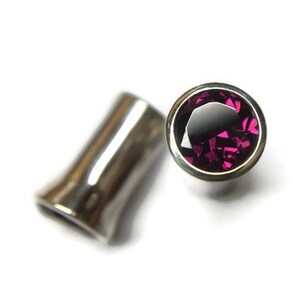 May include: A pair of silver ear gauges, one with a round, faceted, dark pink gemstone set in a silver bezel.
