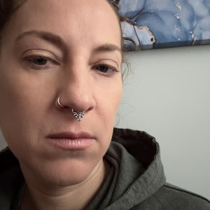 May include: A close-up of a person's face with a silver nose ring featuring a bat design.