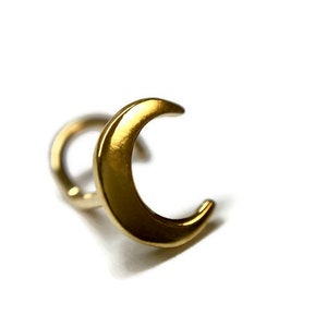 Puede incluir: Un pendiente en forma de media luna de color dorado. El pendiente es de color dorado metálico brillante y tiene una superficie lisa. El pendiente está sobre un fondo blanco.