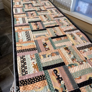 May include: A rectangular patchwork table runner featuring a variety of patterned fabrics in shades of peach, black, and green. The design includes stripes, floral prints, and geometric patterns. The runner is bordered with a black fabric and includes the text "J Cooper Creates Jennifer Cooper".