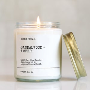 Pu&ograve; includere: Una candela di cera di soia bianca in un barattolo di vetro trasparente con un coperchio dorato. La candela &egrave; etichettata "LULU SUGAR. SANDALWOOD + AMBER 100% Soy Wax Candle Hand poured in Laguna Beach, Calif. scent no. 19"
