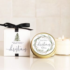 May include: A white square gift box with a black ribbon bow, featuring a green pine tree illustration and the text "HAVE YOURSELF A MERRY LITTLE christmas". Beside it, a candle in a gold tin with a wreath design and the text "The Becks".