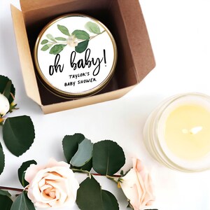 a candle in a box with a label that says "oh baby!" and "Taylor's Baby Shower", along with a candle and some flowers on a white surface.