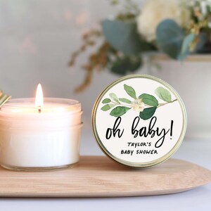 A lit candle in a jar with a label that says "oh baby!" and "Taylor's Baby Shower" written on it, placed on a wooden tray.