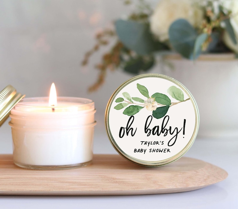 A lit candle in a jar with a label that says "oh baby!" and "Taylor's Baby Shower" written on it, placed on a wooden tray.