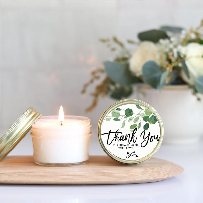 a lit candle in a jar with a "Thank You" label, placed on a wooden surface. In the background, there is a vase with white flowers, creating a serene and elegant atmosphere.