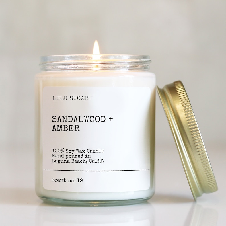 Pu&ograve; includere: Una candela di cera di soia bianca in un barattolo di vetro trasparente con un coperchio dorato. La candela &egrave; etichettata "LULU SUGAR. SANDALWOOD + AMBER 100% Soy Wax Candle Hand poured in Laguna Beach, Calif. scent no. 19"
