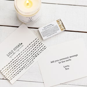 Pu&ograve; includere: Un biglietto bianco con testo nero che recita "LULU SUGAR soy candles + sugar scrubs" e un indirizzo di sito web. Il biglietto &egrave; su una superficie di legno bianco con una candela bianca e una scatola di fiammiferi. I fiammiferi sono etichettati "LULU SUGAR hand poured soy candles" e un indirizzo di sito web.