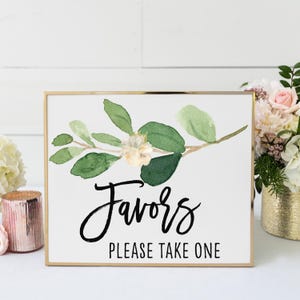 a sign with a floral design and the text "Favorites Please Take One" written on it. The sign is placed on a table, surrounded by flowers and candles.