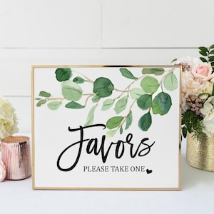 a framed sign with a watercolor design of green leaves and the text "Favorites Please Take One" on it, placed on a table with a vase of flowers and other decorative items.