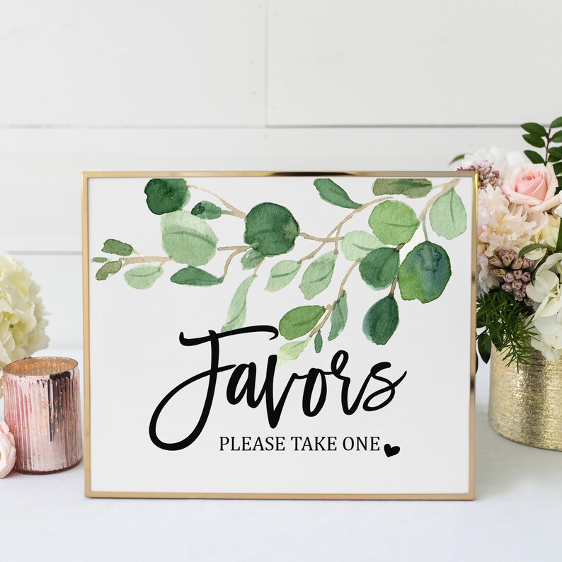 a framed sign with a watercolor design of green leaves and the text "Favorites Please Take One" on it, placed on a table with a vase of flowers and other decorative items.
