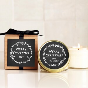 May include: A brown cardboard gift box tied with a black ribbon and a matching candle in a gold-toned tin. Both feature a black square label with a white hand-drawn wreath design and the text Merry Christmas 2024 or love, the sardos.