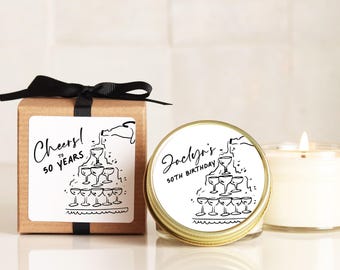 May include: A square brown cardboard box with a black ribbon and a white label featuring a black line drawing of a champagne tower and the text Cheers to 50 Years. Next to it is a round gold tin candle with a matching label reading Jaclyn's 50th Birthday.