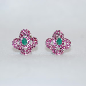 May include: A pair of flower-shaped stud earrings. Each earring has a central oval emerald, encircled by small, round, pink gemstones. The earrings are set against a white background.