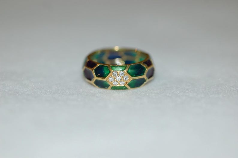 A close-up shot of a gold band ring featuring green and dark enamel in a geometric pattern, centered with a cluster of small diamonds.