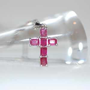 May include: A silver cross pendant with a chain. The cross is adorned with multiple rectangular, vibrant pink gemstones. The pendant is displayed against a blurred, light-colored background, highlighting the jewelry's design and color.