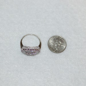 May include: A silver ring with a band of small, clear stones. The ring is shown next to a US dime for size comparison.