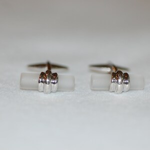 May include: A pair of silver cufflinks with white rectangular stones. The stones have a textured surface and are surrounded by a silver band.