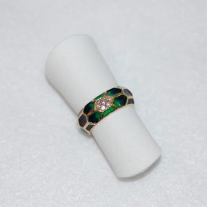 The gold band ring with green and dark enamel and diamond accents is displayed upright on a white cylindrical ring holder against a light background.