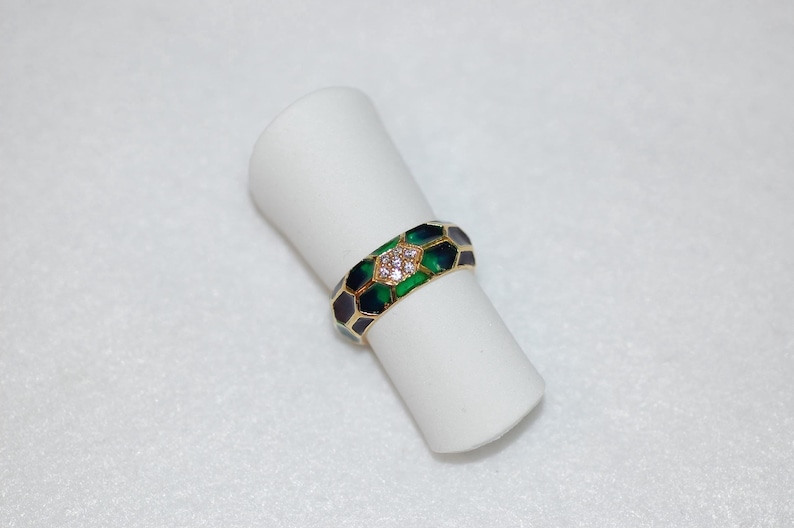 The gold band ring with green and dark enamel and diamond accents is displayed upright on a white cylindrical ring holder against a light background.