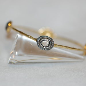 May include: A gold bangle bracelet with a single, large, round, clear diamond surrounded by smaller black diamonds. The bracelet is on a clear display stand.