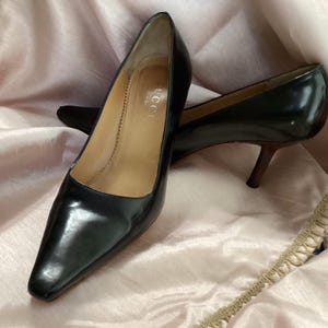 May include: A pair of black leather pumps with a pointed toe and a slim brown heel. The interior of the shoe features a tan leather insole with the Gucci brand name printed in gold lettering. The shoes have a smooth, polished finish throughout.