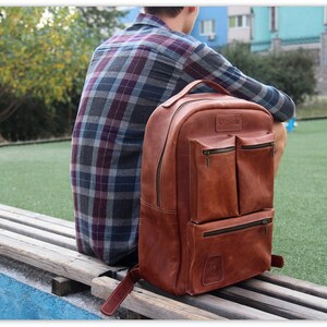 Puede incluir: Una mochila de cuero marr&oacute;n con varios bolsillos y cierre de cremallera. La mochila est&aacute; sentada en un banco de madera.