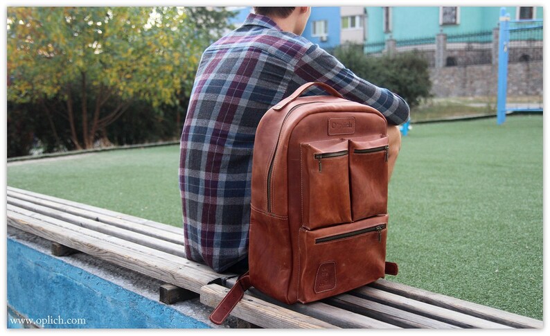 Puede incluir: Una mochila de cuero marr&oacute;n con varios bolsillos y cierre de cremallera. La mochila est&aacute; sentada en un banco de madera.