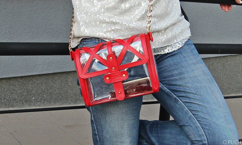 May include: A red and clear plastic crossbody bag with a gold chain strap. The bag has a geometric design and is worn over a denim outfit.