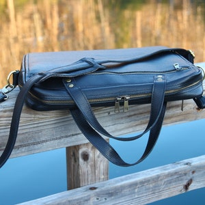 May include: A black leather briefcase with a long strap and two zippered compartments. The bag is resting on a wooden railing.