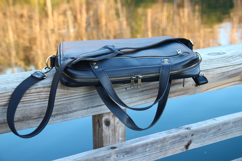 May include: A black leather briefcase with a long strap and two zippered compartments. The bag is resting on a wooden railing.