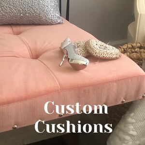 Puede incluir: Un banco de terciopelo rosa con cojines capiton&eacute; y ribete de tachuelas plateadas. El banco tiene un zapato de tac&oacute;n alto plateado y dos posavasos de coraz&oacute;n de ganchillo en la parte superior. El texto "Custom Cushions" est&aacute; impreso en el banco.