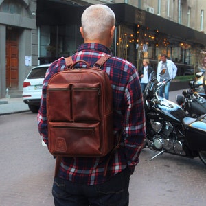 Puede incluir: Un hombre con una camisa de cuadros rojos y negros lleva una mochila de cuero marr&oacute;n con varios bolsillos. La mochila tiene una cremallera y correas ajustables. El hombre camina por una calle de la ciudad.