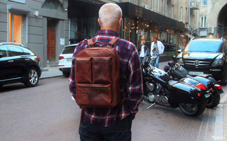 Puede incluir: Un hombre con una camisa de cuadros rojos y negros lleva una mochila de cuero marr&oacute;n con varios bolsillos. La mochila tiene una cremallera y correas ajustables. El hombre camina por una calle de la ciudad.
