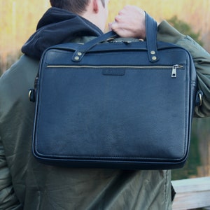 May include: A black leather briefcase with gold hardware and a zipper closure. The briefcase is being carried by a person wearing a green jacket.