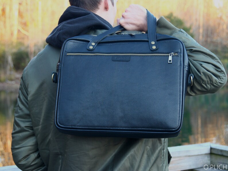 May include: A black leather briefcase with gold hardware and a zipper closure. The briefcase is being carried by a person wearing a green jacket.