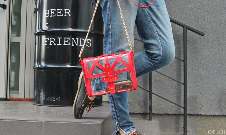 May include: A clear plastic purse with a red trim and gold accents. The purse is worn over the shoulder with a gold chain strap. The purse is worn by a person wearing blue jeans.