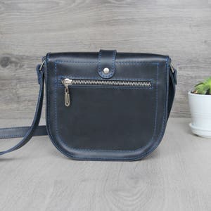Puede incluir: Un bolso bandolera de cuero azul marino con cremallera plateada y un peque&ntilde;o dije plateado. El bolso tiene costuras azules y una correa larga y ajustable. A la derecha del bolso hay una peque&ntilde;a planta en maceta blanca.
