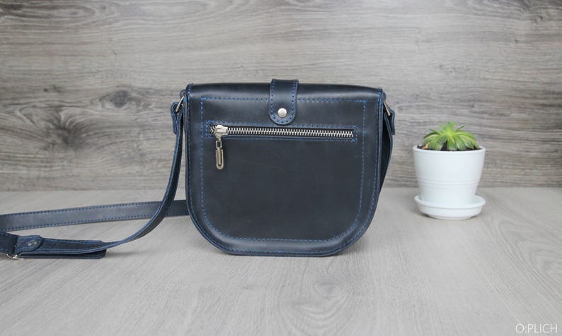 Puede incluir: Un bolso bandolera de cuero azul marino con cremallera plateada y un peque&ntilde;o dije plateado. El bolso tiene costuras azules y una correa larga y ajustable. A la derecha del bolso hay una peque&ntilde;a planta en maceta blanca.