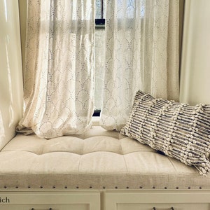 Puede incluir: Un asiento de ventana blanco con un coj&iacute;n acolchado y un coj&iacute;n blanco con un patr&oacute;n texturizado. El asiento de ventana est&aacute; frente a una ventana con cortinas blancas.