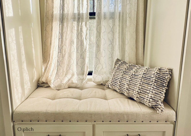 Puede incluir: Un asiento de ventana blanco con un coj&iacute;n acolchado y un coj&iacute;n blanco con un patr&oacute;n texturizado. El asiento de ventana est&aacute; frente a una ventana con cortinas blancas.