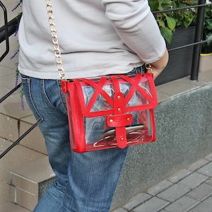 May include: A red clear plastic crossbody bag with a gold chain strap and gold hardware. The bag is worn over the shoulder of a person wearing blue jeans.