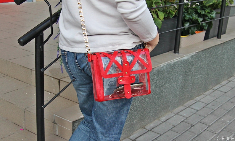 May include: A red clear plastic crossbody bag with a gold chain strap and gold hardware. The bag is worn over the shoulder of a person wearing blue jeans.