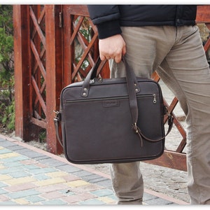 May include: A brown leather briefcase with a zippered pocket and a shoulder strap. The briefcase is being held by a person.