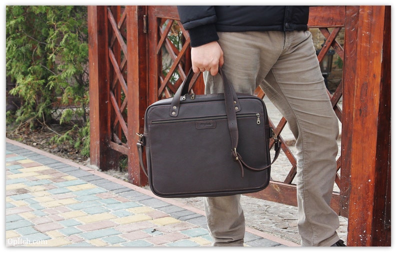 May include: A brown leather briefcase with a zippered pocket and a shoulder strap. The briefcase is being held by a person.