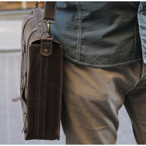 K&ouml;nnte beinhalten: Eine braune Ledertasche mit einem Trageriemen und Messingbeschl&auml;gen. Die Tasche wird &uuml;ber der Schulter einer Person getragen, die ein blaues Hemd und eine braune Hose tr&auml;gt.
