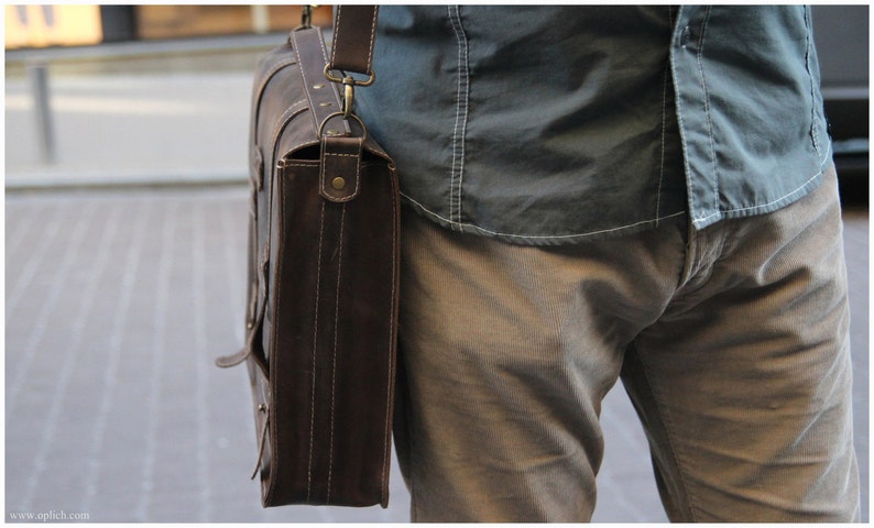 K&ouml;nnte beinhalten: Eine braune Ledertasche mit einem Trageriemen und Messingbeschl&auml;gen. Die Tasche wird &uuml;ber der Schulter einer Person getragen, die ein blaues Hemd und eine braune Hose tr&auml;gt.