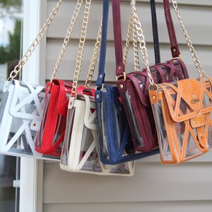 May include: Seven clear crossbody bags with different colored leather accents hanging from gold chains. The bags have a geometric design and are made of clear plastic with a leather flap. The bags are hanging from a white wall.