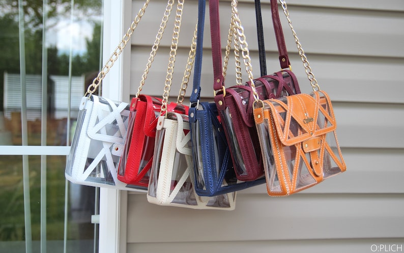 May include: Seven clear crossbody bags with different colored leather accents hanging from gold chains. The bags have a geometric design and are made of clear plastic with a leather flap. The bags are hanging from a white wall.