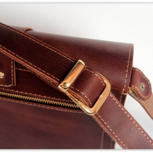 Puede incluir: Primer plano de un bolso mensajero de cuero marr&oacute;n con hebilla y correa doradas. El bolso presenta costuras visibles y una cremallera. La correa est&aacute; sujeta al bolso con una hebilla met&aacute;lica dorada.